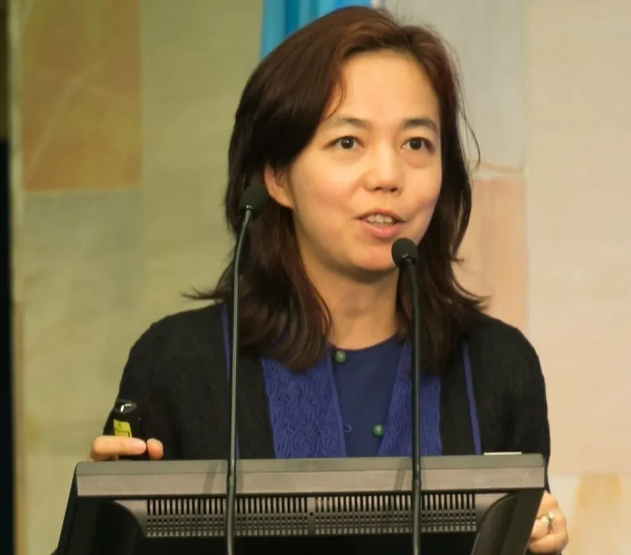 Fei-Fei Li speaking at a podium with two microphones, holding a presentation clicker