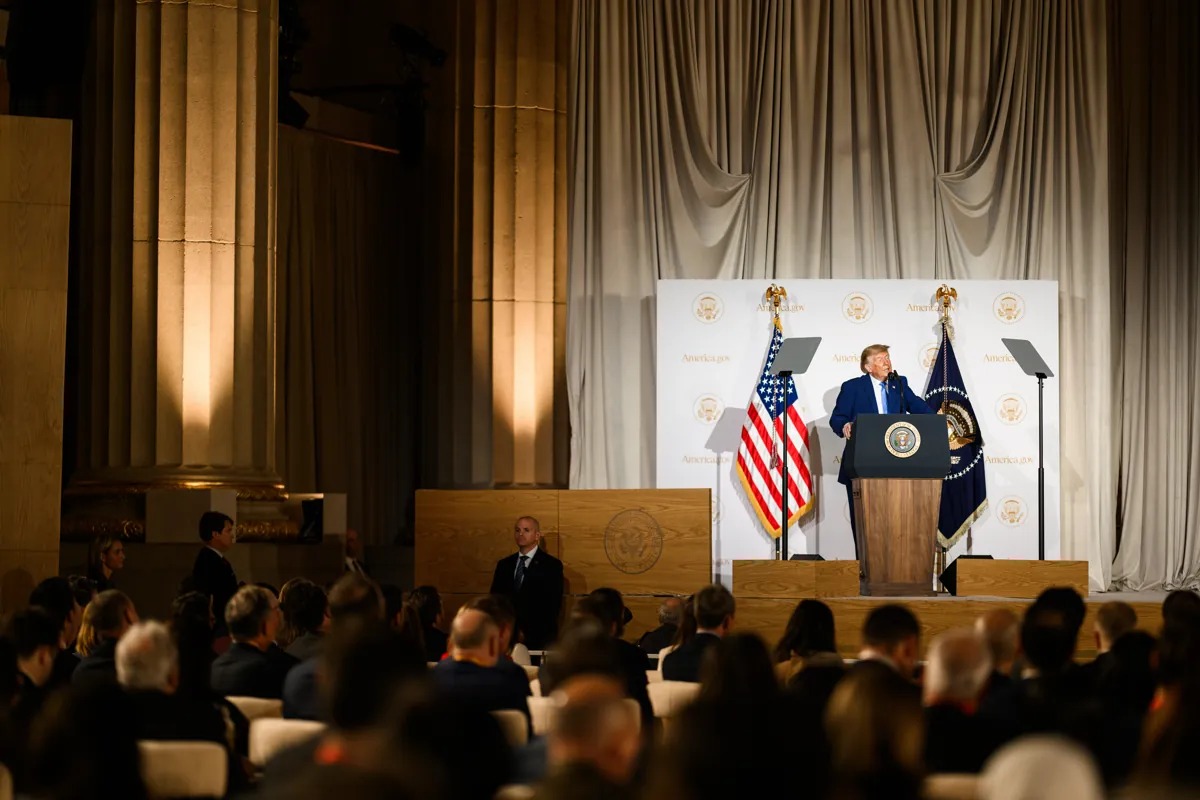 El presidente Donald J. Trump habla en un podio frente a un fondo con el logotipo de America.gov en el Andrew W. Mellon Auditorium, visto desde detrás del público sentado