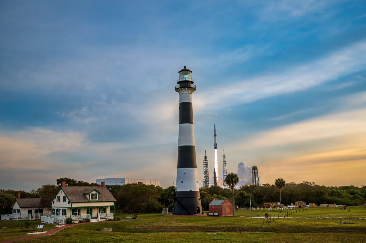 A New Glenn rocket lifting off at dusk behind the black and white Cape Canaveral lighthouse