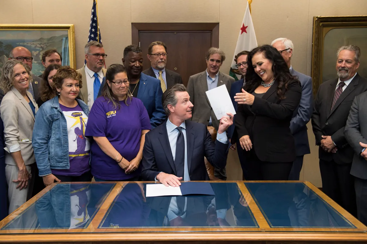 Gavin Newsom seated at a glass-topped desk holding up a signed bill, surrounded by lawmakers and union members in purple shirts