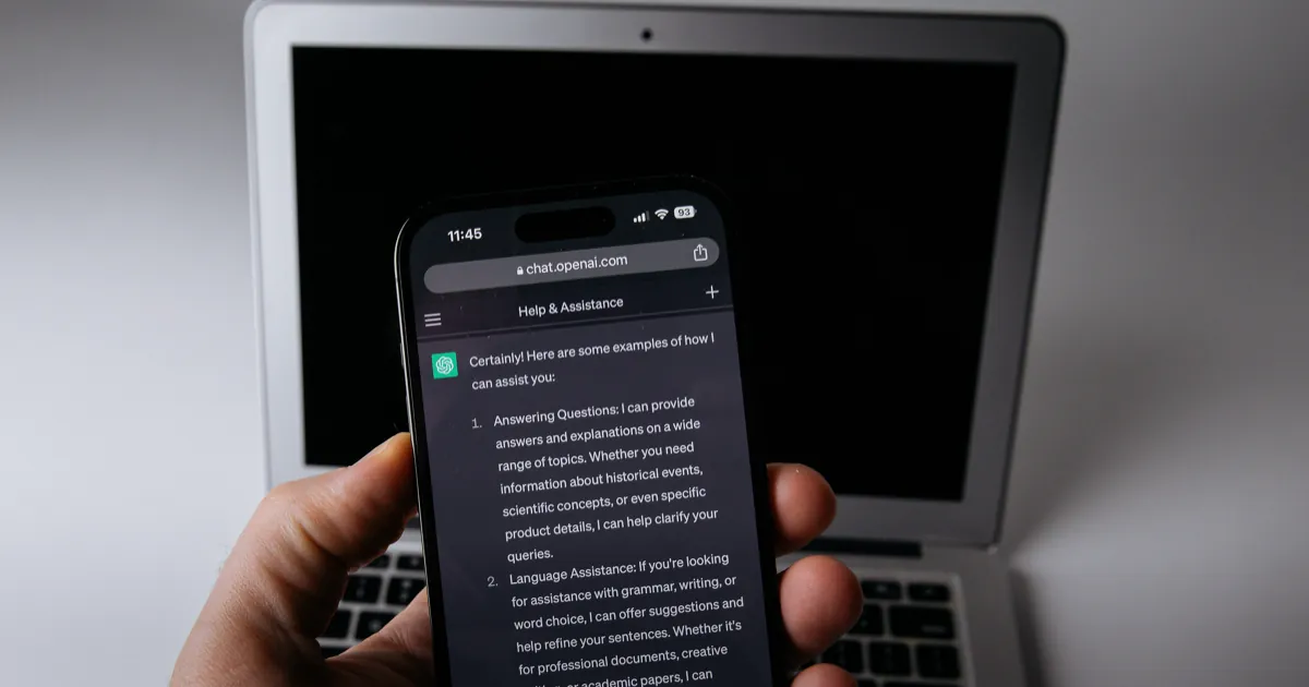 A hand holding a phone with a ChatGPT conversation open in front of a laptop