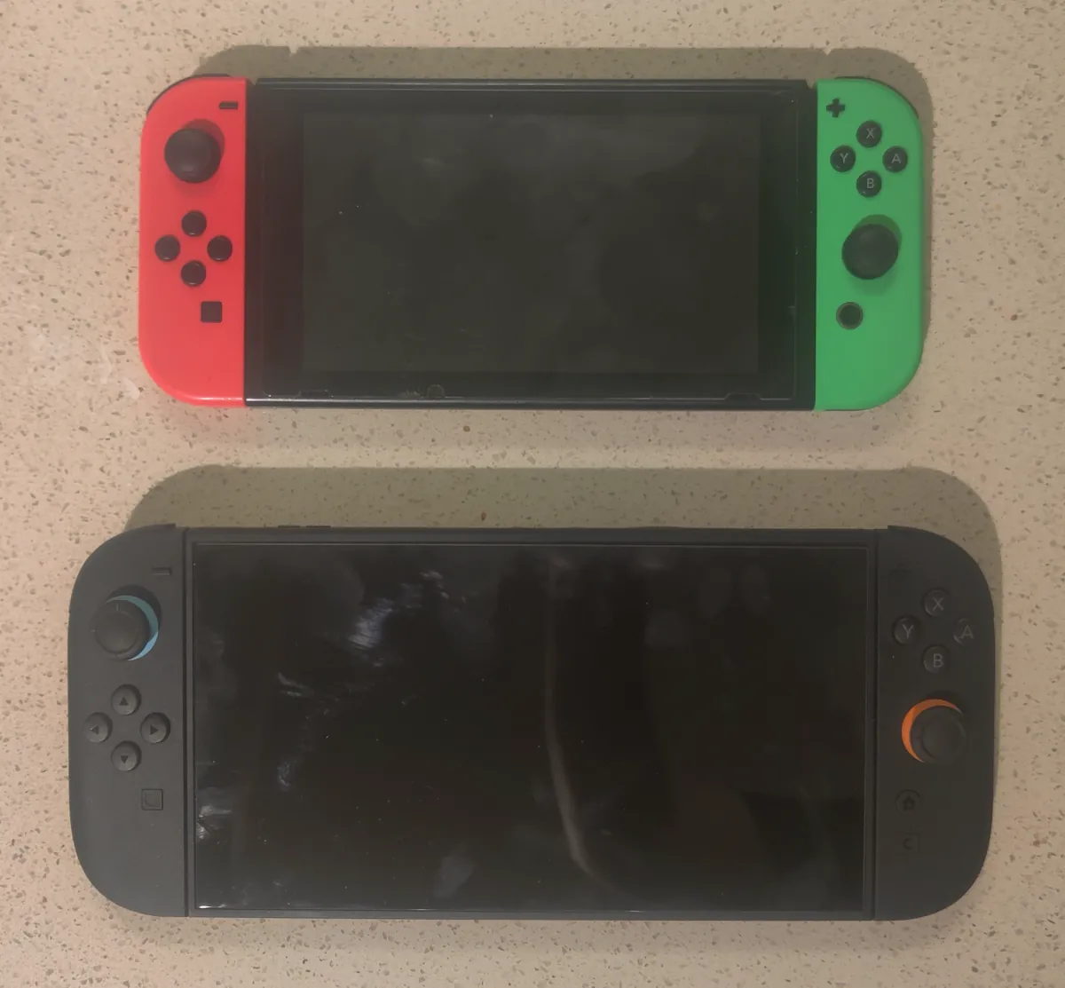 An original Nintendo Switch with red and green Joy-Con above a larger, dark gray Nintendo Switch 2 on a stone countertop