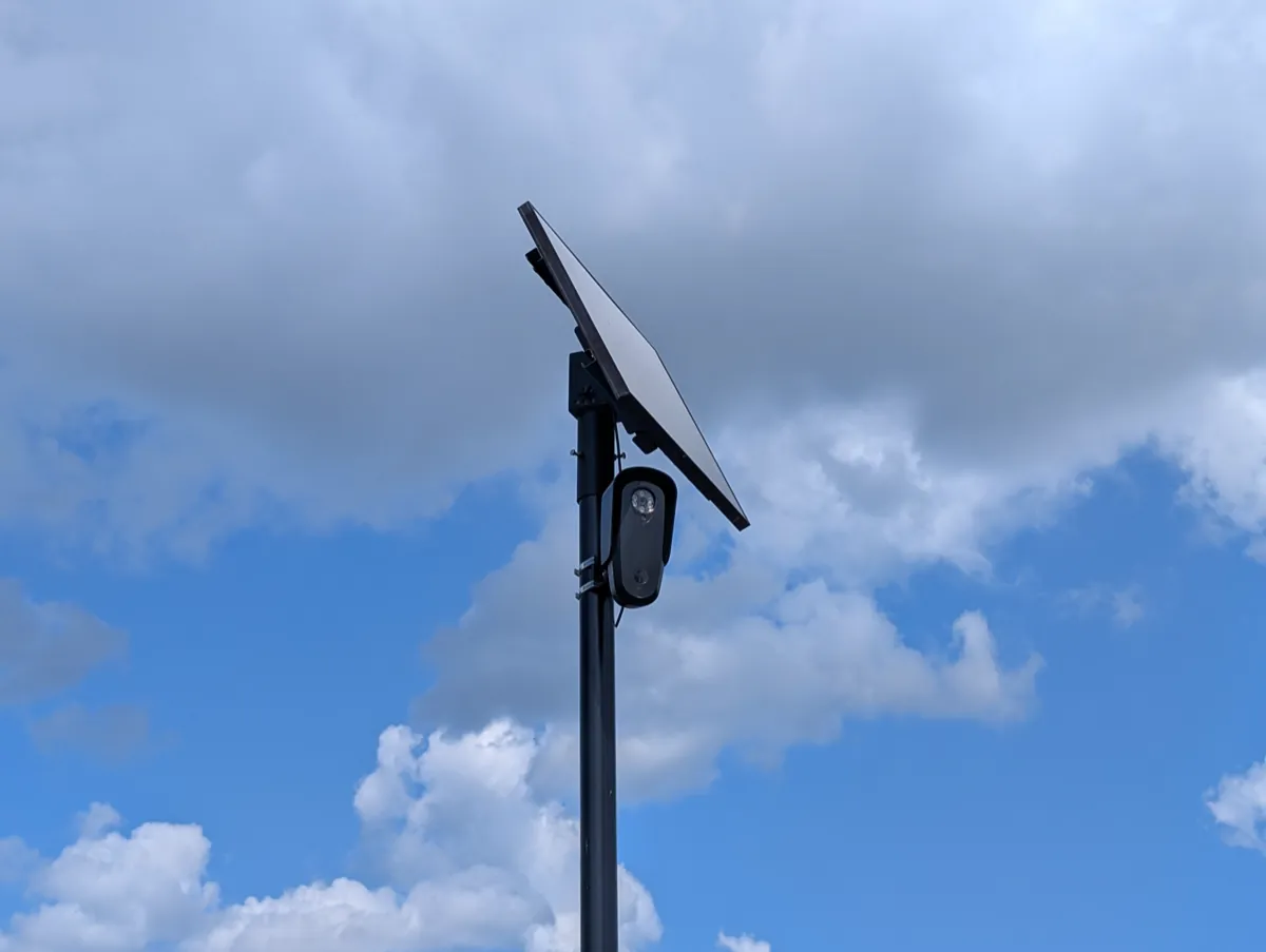 A Flock license plate reader camera and its solar panel on a tall black pole against a blue sky with clouds