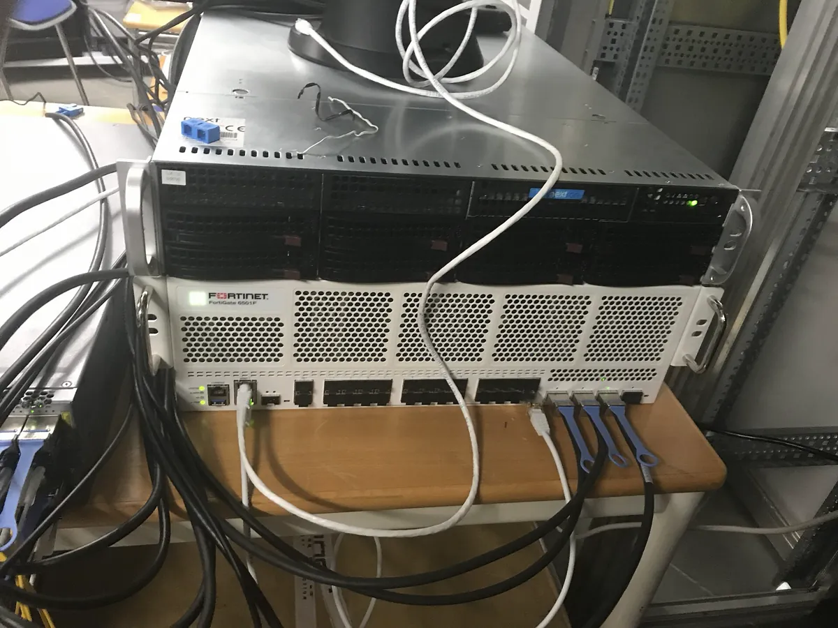 A white Fortinet FortiGate 6501F firewall mounted below a black server in a rack, with network cables plugged into its front ports