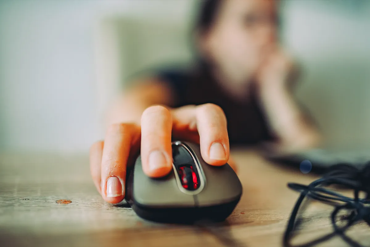 Close-up of a hand resting on a dark computer mouse on a wooden desk, with a person blurred in the background