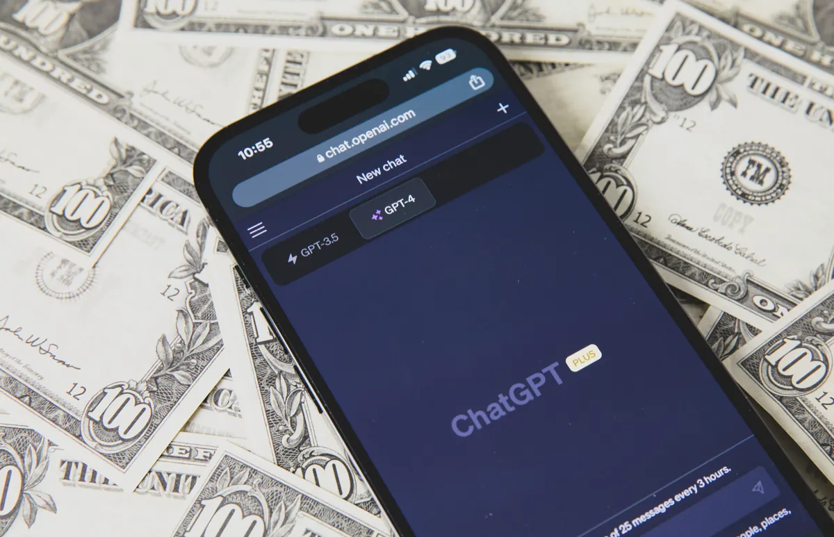 A phone showing an older ChatGPT Plus screen, lying on a spread of prop US$100 bills