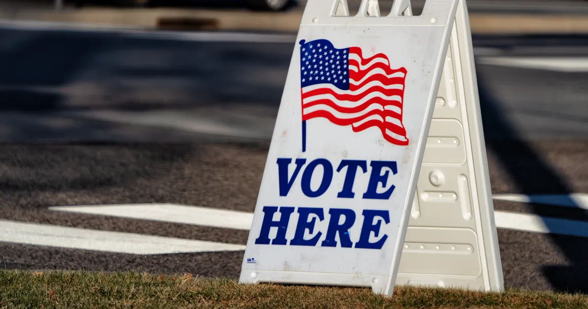 A white Vote Here sign with an American flag standing on the grass outside a polling place