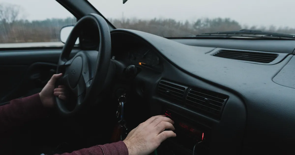 A driver's hand turning the dial of a car radio on a dark dashboard, with a gray winter road outside the windshield