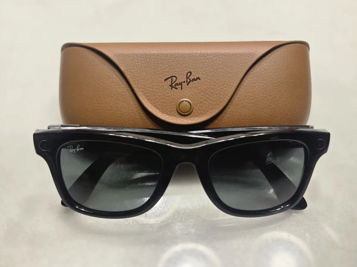Black Ray-Ban Meta Wayfarer smart glasses in front of their brown leather case, with a small round camera and light at each corner of the frame
