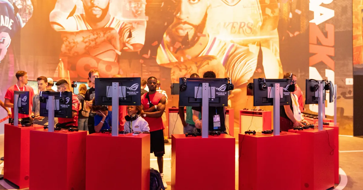 Players at red NBA 2K20 demo stations at Gamescom 2019, under a giant mural of NBA stars