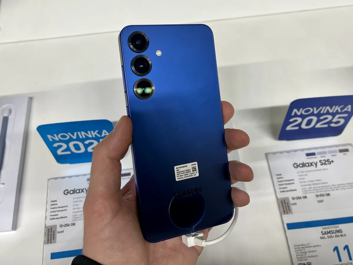 A hand holding a blue Samsung Galaxy S25+ from the back in a store display, with its three rear cameras visible