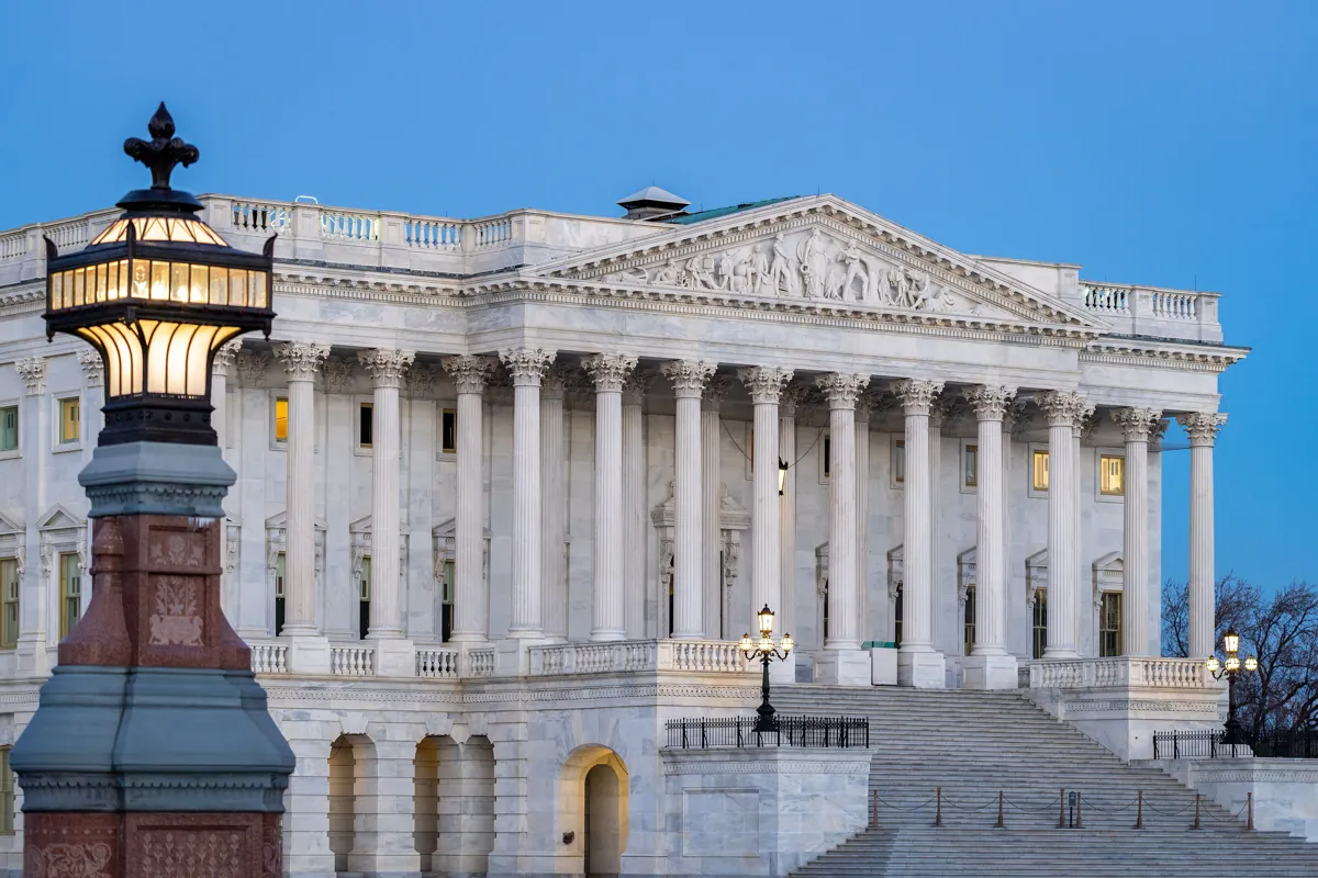 The Senate wing of the US Capitol at dusk, with marble columns and a lit lamp in the foreground