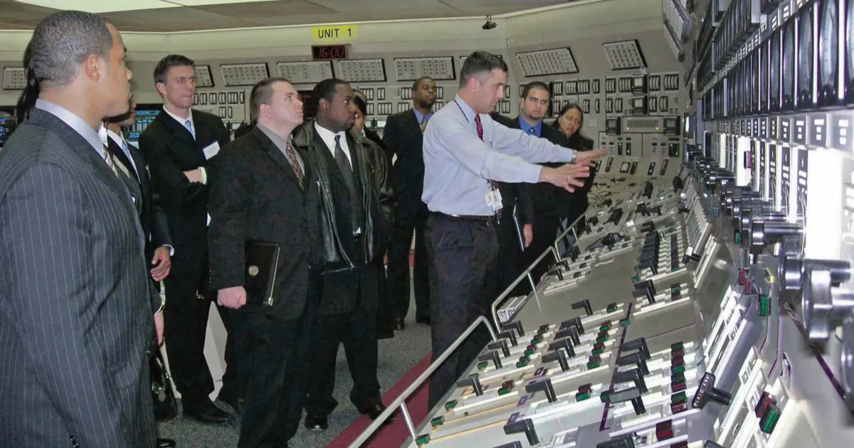 A guide points at the panels of a nuclear power plant control room while a group of visitors in suits looks on