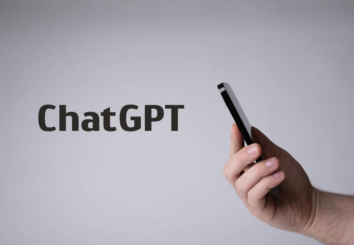 A hand holding a phone next to the word ChatGPT printed on a gray background