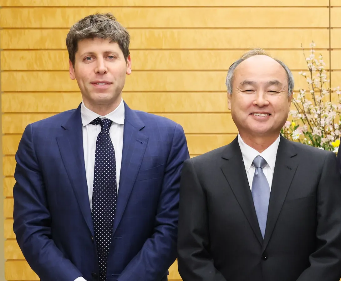 Sam Altman in a navy suit and polka-dot tie standing next to a smiling Masayoshi Son in a dark suit, in front of a wooden wall