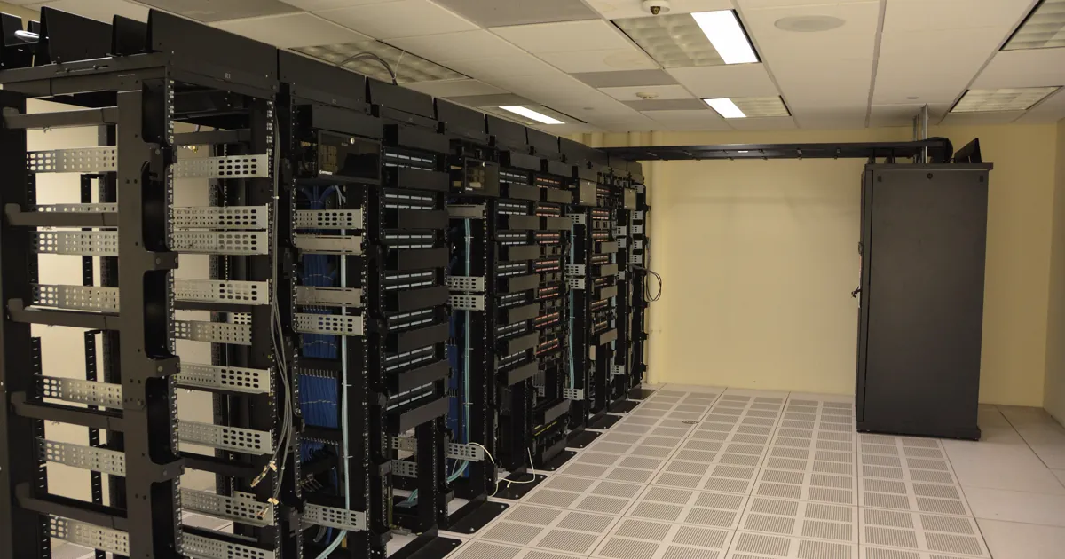 Rows of black server racks with blue network cabling in a data center room with a raised tile floor