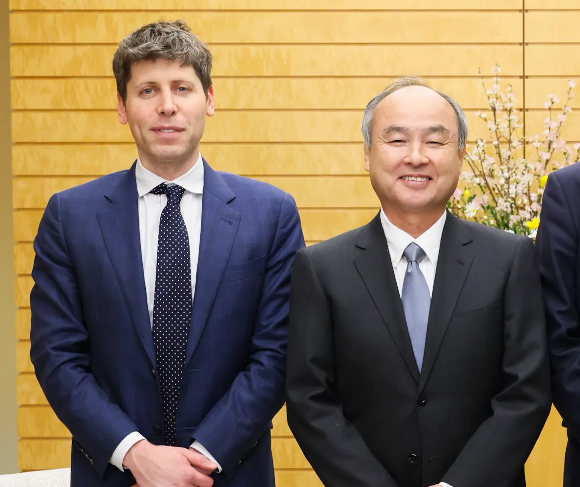 OpenAI CEO Sam Altman in a navy suit and polka-dot tie standing beside a smiling Masayoshi Son in a dark suit, in front of a wooden wall