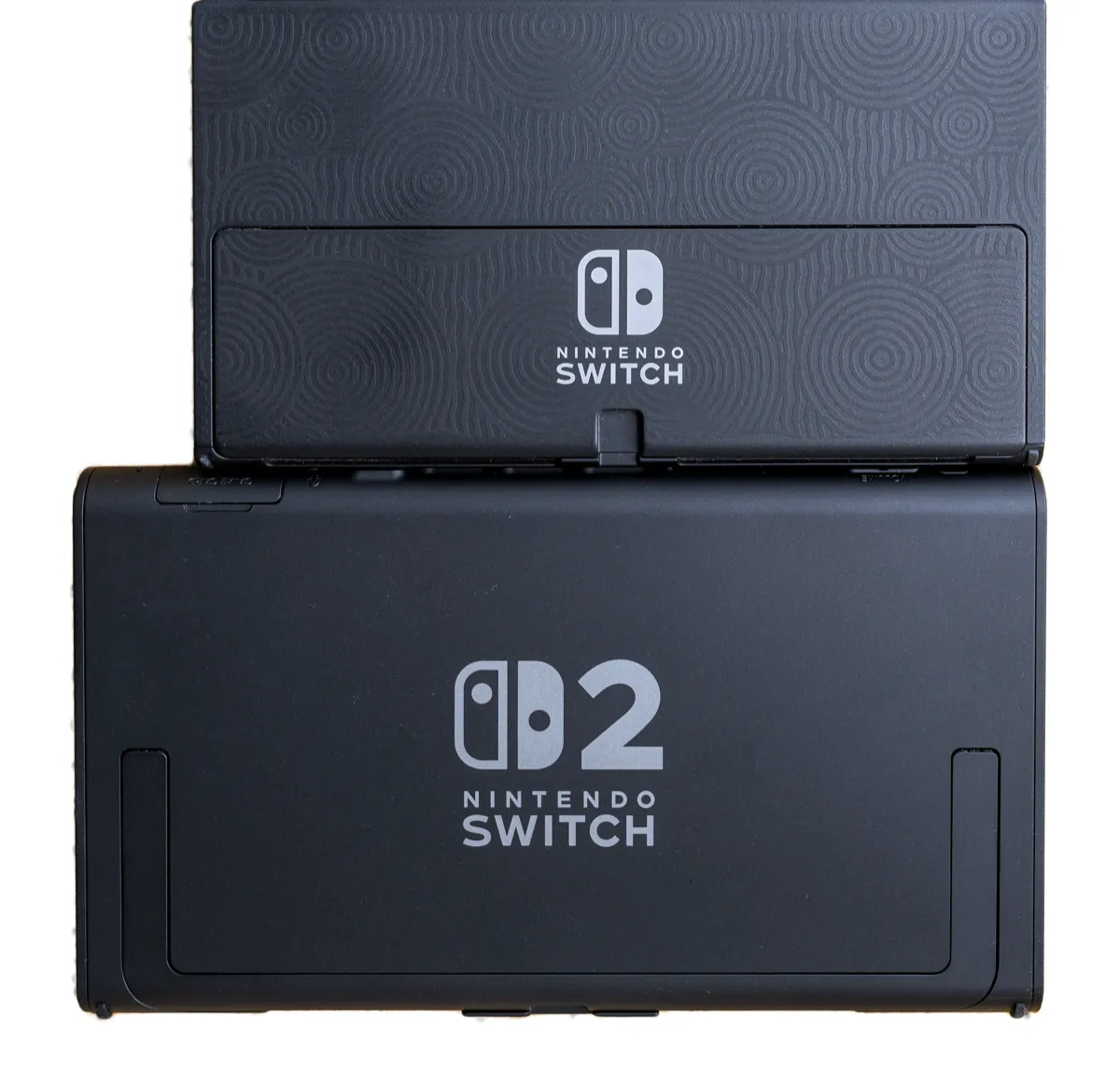 The back of a Nintendo Switch OLED Model stacked above the larger back of a Nintendo Switch 2, both showing their logos
