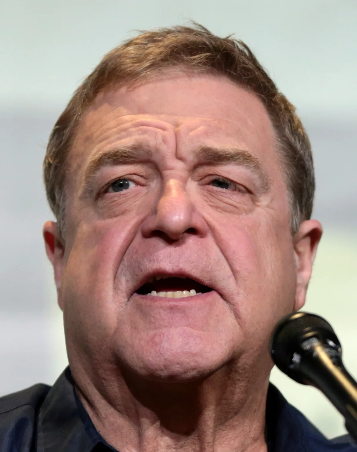 John Goodman speaking into a microphone on a convention panel in 2016