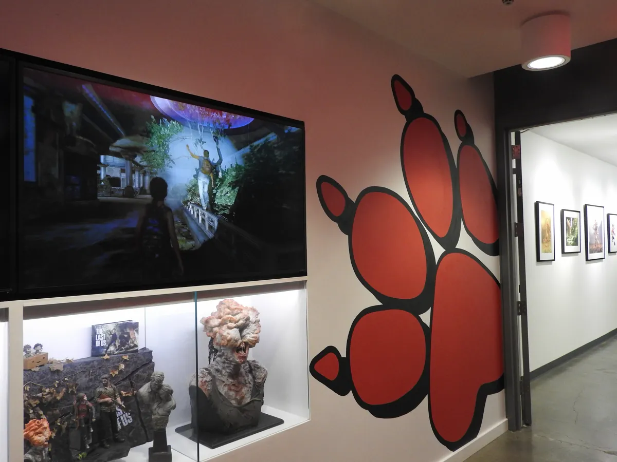 Hallway at Naughty Dog's studio with a red paw print mural, a screen and The Last of Us statues in a display case