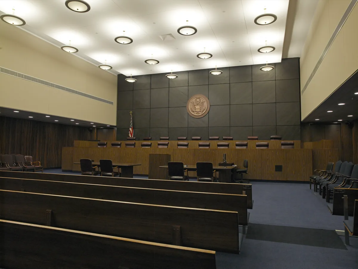 An empty courtroom with a long two-tier judges' bench, wooden pews and the US seal on a dark stone wall