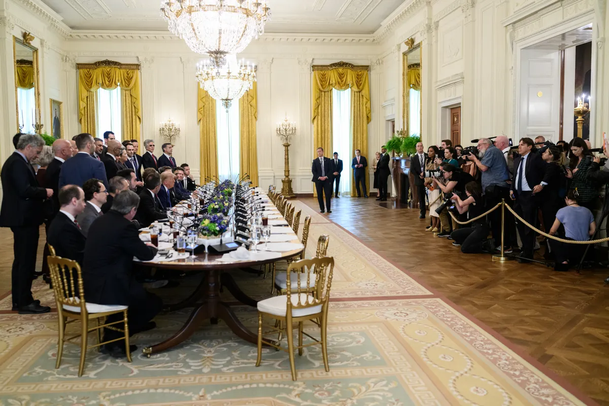 Guests seated along a long lunch table in the White House East Room under crystal chandeliers, with press photographers behind a rope line