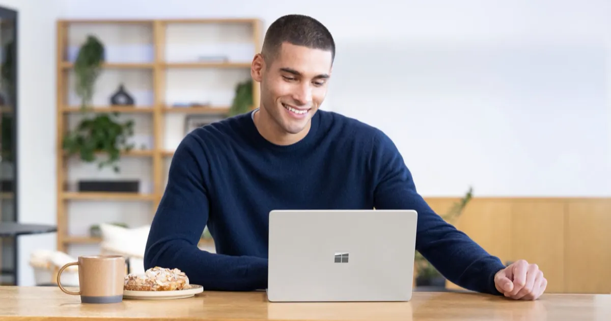 A man smiling at a silver Microsoft Surface laptop on a wooden table, next to a coffee mug and a pastry