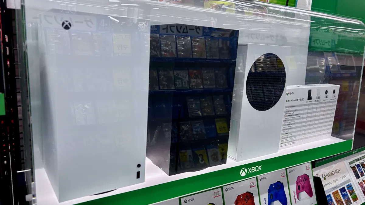 White Xbox Series X and Xbox Series S consoles on a lit store shelf above colored Xbox controllers and Game Pass cards
